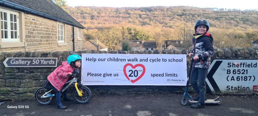 A photograph showing 2 children, each side of a banner that says 'help children walk and cycle to school please give us 20mph speed limits' One child is on a balance bike, the other a scooter. Both wear helmets and a wide grin.
